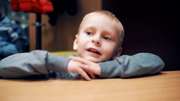 部屋のテーブルに座ってカメラを見ている男の子の肖像画 面白い話と彼の小さな顔に異なる感情を作る青い目の好奇心旺盛な子供 — ストック動画