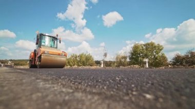Turuncu paten makinesi yazın yolda sıcak asfalta basıyor. Ağır sıkıştırma makinesi mavi gökyüzü ve yeşil ağaçların arka planında yeni asfalt oluşturuyor. Yol yüzeyi.