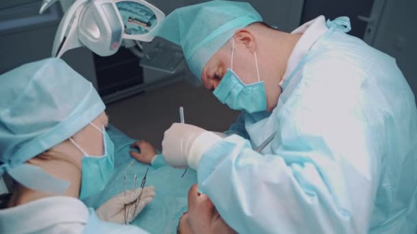 歯医者の男は歯科医院で患者の口の中で糸で縫っている 室内で患者さんと一緒に働く専門家の男性と彼のアシスタント 歯科手術 — ストック動画