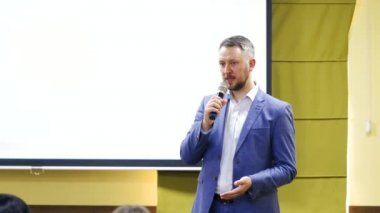 İş adamı erkek, konferans sırasında yüzünde bir gülümsemeyle bir konferans veriyor. Beyaz ekranın arka planında konuşan şık mavi takımlı bir adam..
