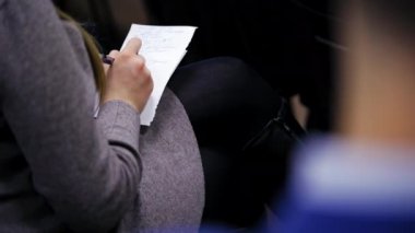 Genç bayan konferans sırasında bir kağıt parçasına bilgi yazar. Kadın, uluslararası kongrede elinde kalem ve defterle bir sandalyede oturuyor..