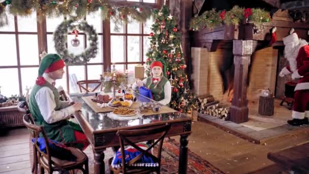 圣诞老人和两个精灵在准备茶 圣诞老人和沸腾的茶壶 好奇的男孩和女孩穿着精灵的衣服 穿着童装的快乐精灵坐在桌旁 圣诞壁炉附近的圣诞老人 — 图库视频影像