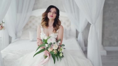 Seksi gelinlikli çekici genç gelin, yatakta oturuyor. Çiçekli güzel gelin. Beyaz elbiseli güzel manken yatak odasındaki kadın fotoğrafçıya poz veriyor..