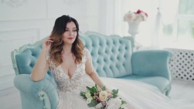 Evde çiçekli bir kanepede oturan gelinlik içindeki göz kamaştırıcı gelin. Güzel dantelli bir elbise ve şık bir stüdyo arka planında poz veren modern saç modeli..