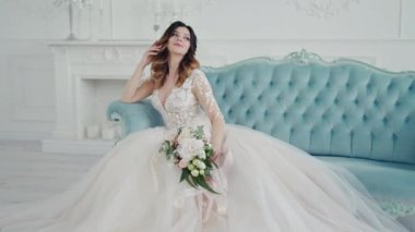 Kanepede oturan beyaz gelinlikli inanılmaz genç bir kadın var. Güzel bir model. Makyajlı. Kraliyet kanepesinde poz veriyor ve kameraya gülümsüyor..