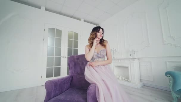 結婚式のドレスの美しい花嫁は部屋の椅子に座っている間彼自身を賞賛する きれいなドレスの柔らかいモデルは 紫色のアームチェアの上に光スタジオでポーズをとっています — ストック動画
