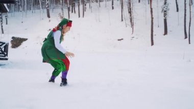 Yeşil takım elbiseli genç elf kızı ve karda neşeyle koşan bir şapka. İki elf arkadaş kışın ormanın arka planında birlikte kartopu oynuyorlar. Yavaş çekim.