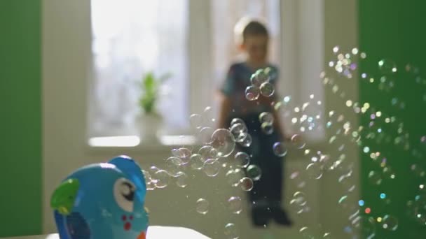 男の子と一緒にぼやけた背景の部屋にカラフルな円の泡が飛んでいます テーブルの上に石鹸泡を作成するための特別なデバイス 部屋の中のシャボン玉 — ストック動画