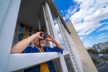 Genç bir çocuk balkondan dürbünle bakıyor. Mavi gökyüzü ve şehir manzarası arka planı.