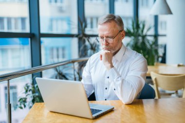 Beyaz gömlekli, düşünceli, orta yaşlı bir iş adamı. Pencerenin yanında dizüstü bilgisayarı olan bir adam çalışıyor. İş konsepti.