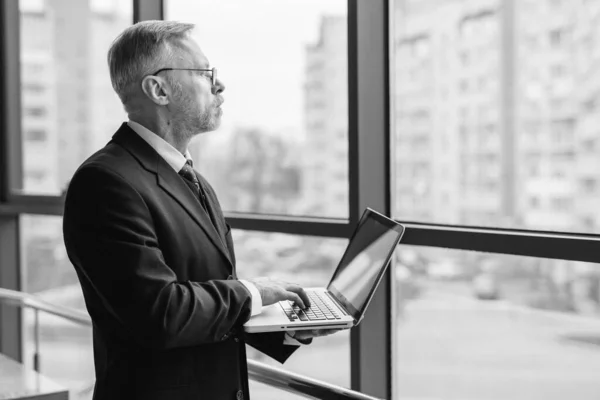 Bearded man standing near window with laptop. Panoramic city view. Black and white photo.