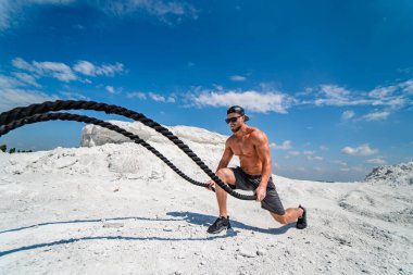 Güçlü vücutçular kaslarını şişiriyor. İplerle antrenman. Vücut geliştirme ve açık hava sporları konsepti. Açık havada poz vermek. Taş ocağında antrenman. Yan görüş. Beyaz manzara.