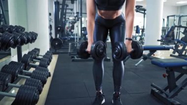 Spor salonundaki metal halterlerle egzersiz yaparken seksi bir kadın vücudu. Vücut geliştirici kadın dambılları kaldırıyor ve aynaya bakıyor fitness kulübünün arka planındaki yansımasında..