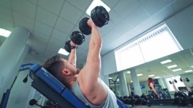 Kolları masanın üzerinde duran ve spor salonunda dambılları kaldıran güçlü bir adamın yan görüntüsü. Kaslı adam spor merkezinin arka planında egzersiz yapıyor. Güzel kadın vücut geliştirme eğitimine bakıyor..