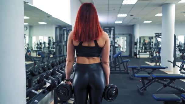 体育館でのスポーツウェアリフティングダンベルで完璧なボディを持つアスレチック若い女性 赤い髪の女性はスポーツクラブの背景に訓練中にダンベルで彼女の筋肉をやっています — ストック動画