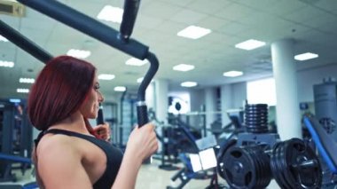 Güzel güçlü kadın vücut geliştirici spor salonunda kaslarını geliştirmek için egzersiz yapıyor. Spor kıyafetli, kaslı, kızıl saçlı kız modern bir simülatörde egzersiz yapıyor..