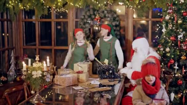 小さな子供がサンタの家からXmasの背景に封筒を取っています テーブルにタイプしているサンタクロースの近くで踊っている妖精の衣装の幸せなエルフ 内部のクリスマスの雰囲気 — ストック動画