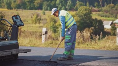 Asfalt makinesi otoyolda asfaltı gündüz ağır makinelerin arka planında bırakır. Koruyucu üniformalı işçiler yol inşaatı sırasında asfalt kaldırım makinesinin çalışmasını kontrol ediyorlar.