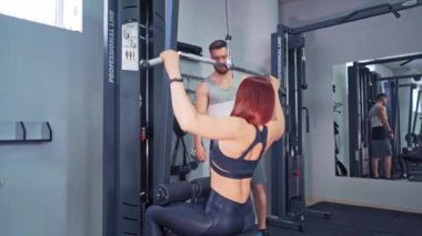 Israrcı kadın idmanını simülatörde yapıyor. Atletik kadın spor salonunda kendine bir çubuk çekerek kaslarını çalıştırdı. Profesyonel antrenör, sporcunun iç mekandaki eğitimine bakıyor..