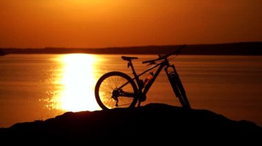 Günbatımının arka planında modern bir bisiklet. Nehir kenarında bisiklet ve gece vakti kask ve spor giysiler giyen bir adamın silueti..