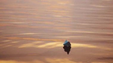 Akşam suyunda kağıt tekne. Beyaz origami teknesi gün batımında su yüzeyinde tek başına yüzüyor..