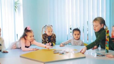 Okulda resimlerle İngilizce öğrenen küçük öğrenciler. Yuvarlak masada oturan ve ışık sınıfındaki kartlara bakan çocukların eğitim sürecini öğretmen kontrol ediyor..