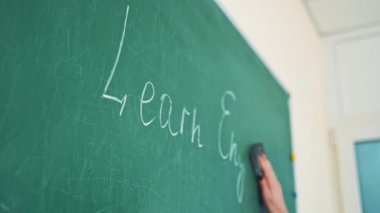 Yakın çekim öğretmeninin eli tahtadaki tebeşiri kaldırıyor. Sınıfta bir süngerle İngilizce yazıları silen bir kadın..