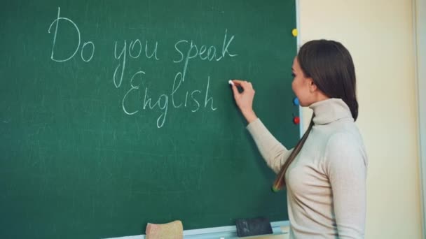 有魅力的英语教师用粉笔在绿色的黑板上写字 美丽的长发女子正在黑板上写问题标签 并在课堂上对着摄像机微笑 — 图库视频影像