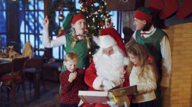 Noel Baba 'nın Noel arkaplanda dans eden küçük çocukları ve elfleri var. Noel Baba çocuklara fotoğraf albümünü gösterirken neşeli elfler evde eğleniyor..