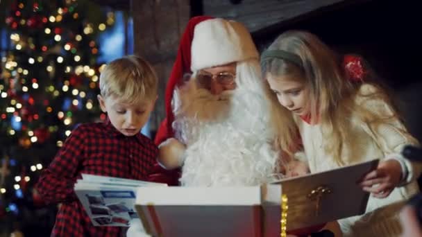 산타클로스와 앨범을 호기심많은 소년과 산타가 크리스마스 트리의 배경에 아이들에게 앨범을 — 비디오