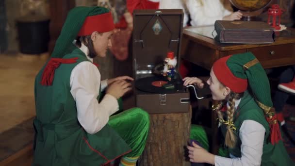 穿着童装的滑稽精灵在屋里玩玩具 圣诞老人的住宅里 两只小精灵和玩具士兵在旧录音机上旋转着一个圆盘 — 图库视频影像