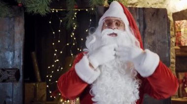 Uzun beyaz sakallı Noel Baba şarkı söylüyor. Noel Baba kırmızı kostüm ve beyaz eldivenler giyip Noel arka planında sakalını düzeltiyor..