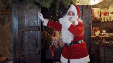 Kırmızı ve beyaz kostümlü Noel Baba çok fazla el kol hareketi yapıyor. Beyaz sakallı Noel Baba Noel hediyelerinin arka planında ellerini sallıyor..