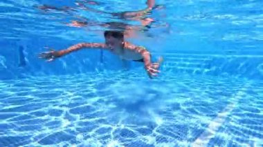 Çocuk yüzme havuzunda suyun altında yüzüyor. Suyun altında ve genç adam açık mavi sularda gözleri kapalı yüzüyor..