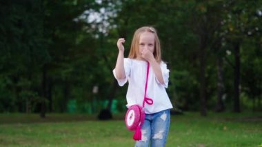 Beyaz bluzlu neşeli sarışın çocuk, küçük pembe çantalı kot, burnunu kapatırken mutlu bir şekilde dans ediyor.