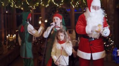 Christmas celebration with Santa. Fairy elves, Santa and children are singing and dancing on Christmas background indoors.