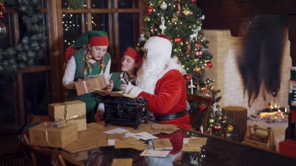 桌上有很多礼物 还有圣诞老人和精灵在一起房间里有圣诞背景精灵和圣诞老人正试图猜测盒子里有什么 — 图库视频影像