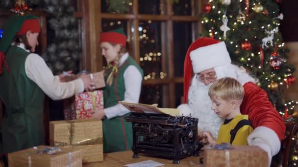 圣诞老人正在教小男孩在一台旧打字机上打字 圣诞节的时候两只身穿绿色服装 盒子里有礼物的精灵 — 图库视频影像