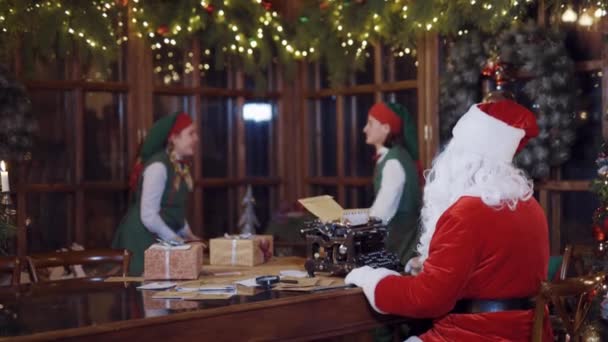 身穿绿色童装的快乐精灵们玩箱子 圣诞老人正在桌上打字 而快乐的精灵们则在圣诞节装饰的背景下享受着礼物的乐趣 — 图库视频影像