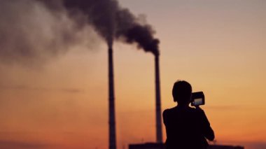 Gün batımında telefonu olan bir adamın silueti. Arka planda siyah dumanlı iki borunun üzerinde akşam gökyüzünün fotoğraflarını çeken bir adam. Ekolojik sorunlar.