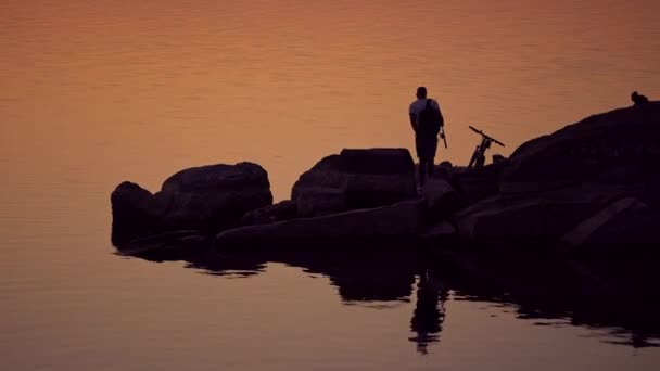 夕方の水の背景の彼のバイクの近くの釣竿を持つ男 リュックサックを持つ漁師のシルエットは 水の上に石の岩の上に立って家を集めています — ストック動画