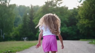 Parkta koşan bir kızın arka görüntüsü. Uzun saçlı mutlu küçük kız yazın doğal yeşil arka planda tek başına eğleniyor..