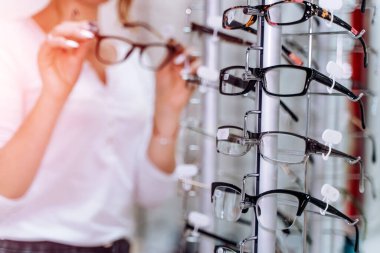 Kadın, arka plandaki Optik Dükkanı 'nda elinde gözlüklerle ayakta duruyor. Gözlükleri takın. Görme düzeltmesi. Kapat..
