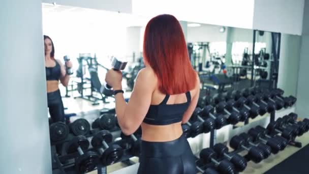 女健美运动员在体育馆接受哑铃训练 红头发女子 肌肉发达 双手托起哑铃 站在镜子前 — 图库视频影像
