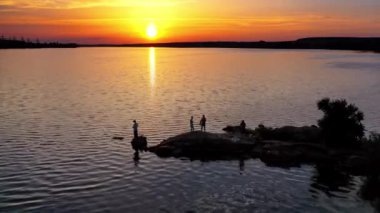 İnsanlar akşamları suyun yanında taşların üzerinde duruyorlar. Nehrin doğal arka planında insansız hava aracı görüntüsü ve gün batımında insan siluetleri. Kamera yukarı hareket ediyor.