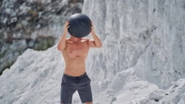 Güneş gözlüklü genç adam vücudunu doğal olarak eğitiyor. Güçlü kaslı sporcu güneşli bir yaz gününde dağlarda topla çömelir..