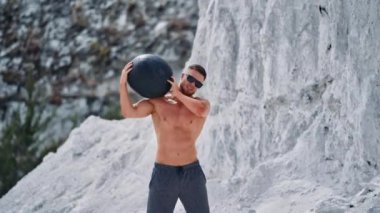 Beyaz dağlarda ağır bir topla vücut geliştirici eğitimi. Siyah güneş gözlüğü takan güçlü bir adam yaz aylarında kafasına top geçirip egzersiz yapıyor..