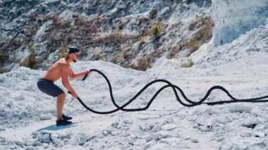 Vücut geliştirme eğitiminin yan görüntüsü, dışarıda dövüş halatları. Yakışıklı sporcu ellerini hızla dağın arka planında kabloyla hareket ettiriyor..