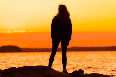 Kayanın üzerinde dik duran bir kadın. Doğa ve güzellik kavramı. Turuncu gün batımı. Gün batımında kız silueti.