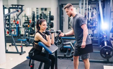 Genç, güzel esmer fitness kızı halter diskiyle spor salonunda antrenman yapıyor. Antrenör, daha iyi sonuçlar için yeniden uzlaşma sağlıyor.